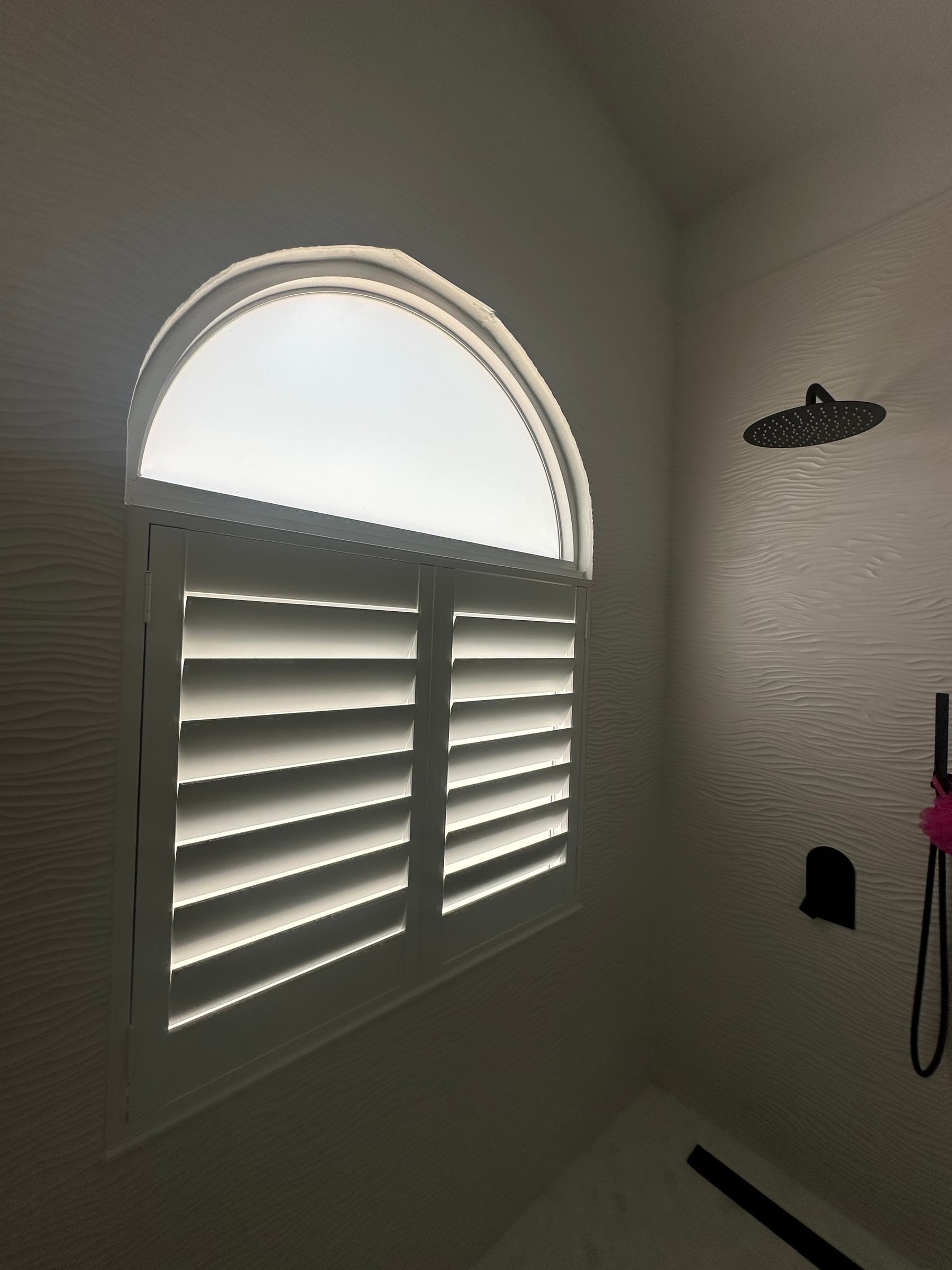 A shutter panel below a fixed arched window in a tiled shower alcove