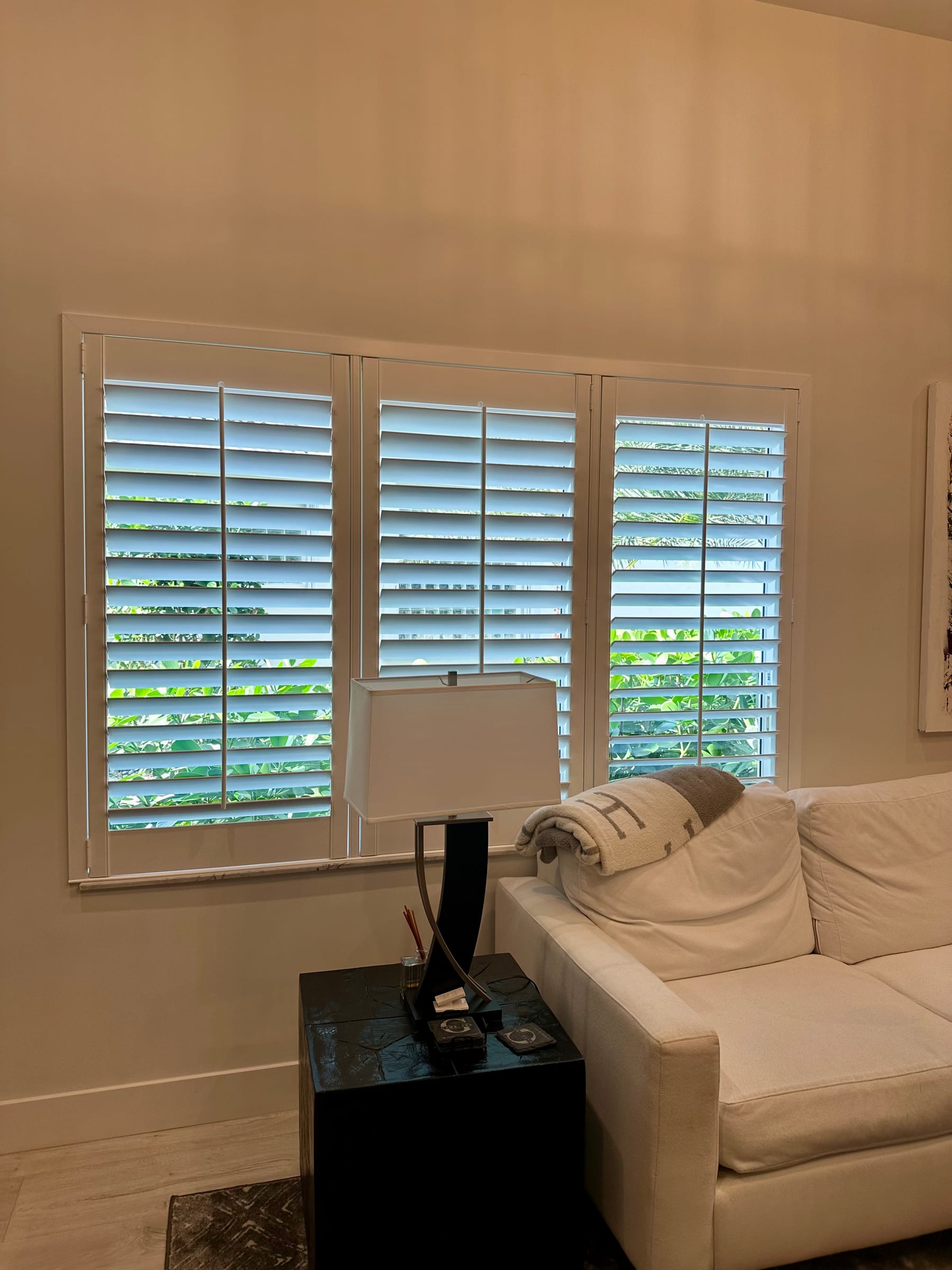 White shutters across a window behind a pale sofa and a table lamp