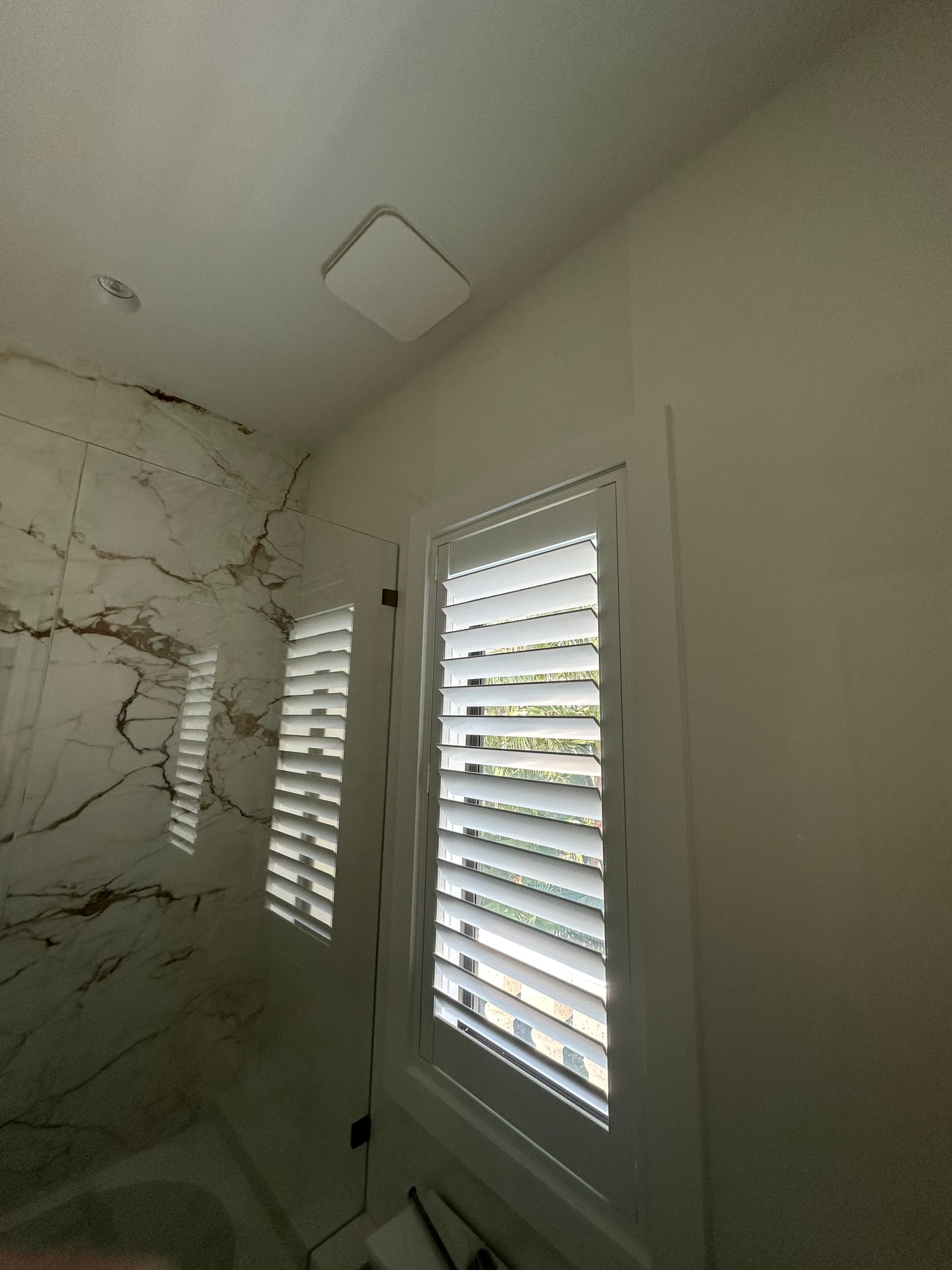 A white shutter in a marble bathroom beside a glass shower screen