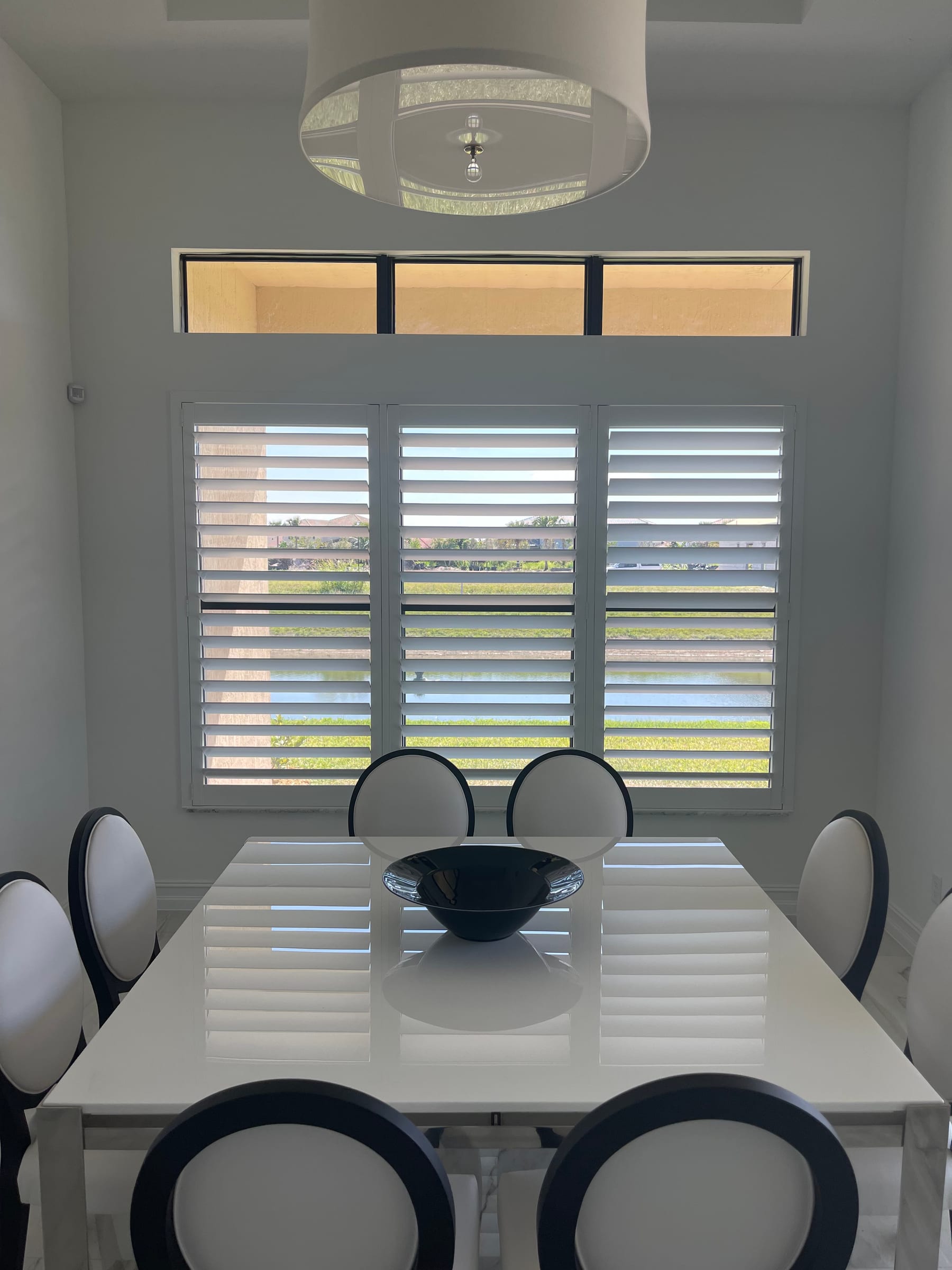 White shutters across a dining-room window wall with clear transoms above