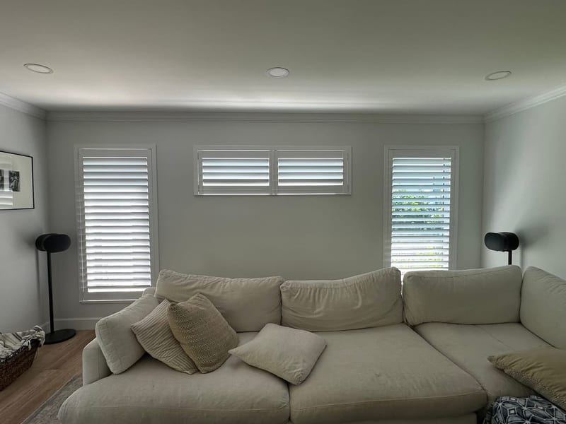 White shutters on the windows around a living-room sofa, louvers open to daylight